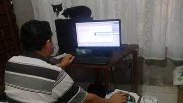 Ramón Darío Marín en un computador de mesa, una gata sentada en la torre del computador